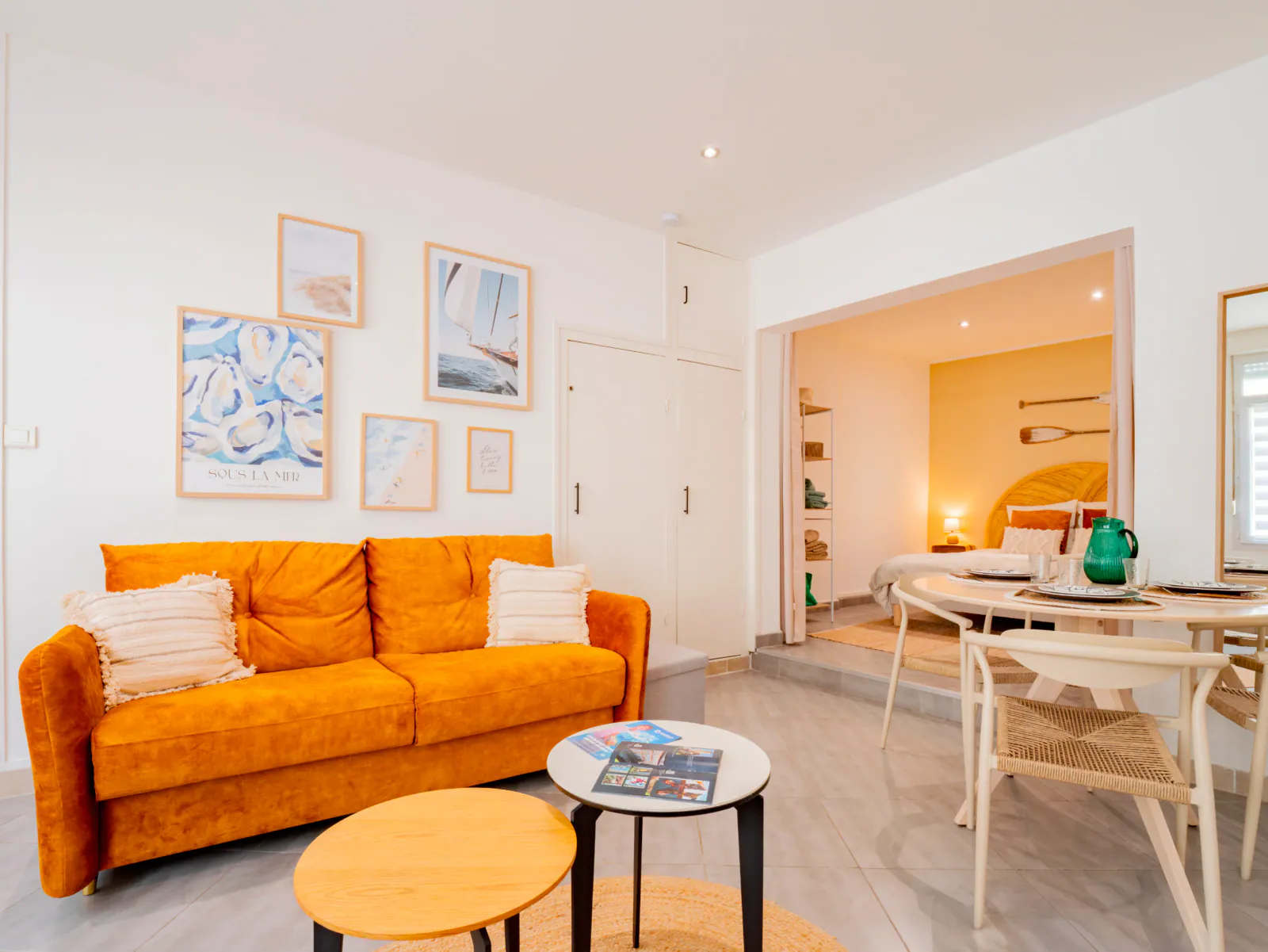 Salon lumineux d'un gîte côtier avec canapé ocre, tableaux marins et vue sur chambre